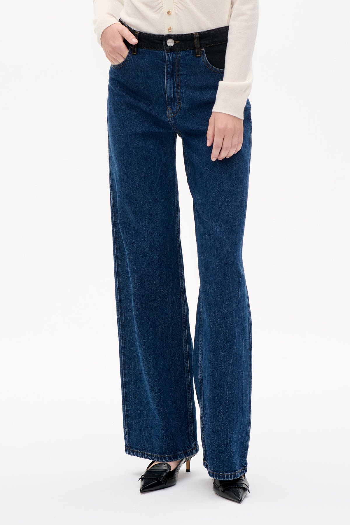 Front detail of dark blue wide-leg jeans with a contrasting black waistband, gold-tone stitching and button, styled with an ivory cardigan.