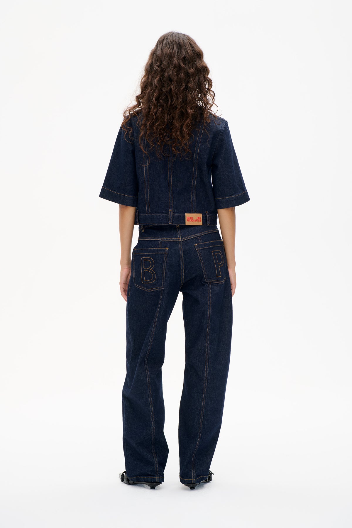 Back view of a dark indigo denim short-sleeved jacket with contrast stitching, paired with matching wide-leg jeans and lettered back pockets.