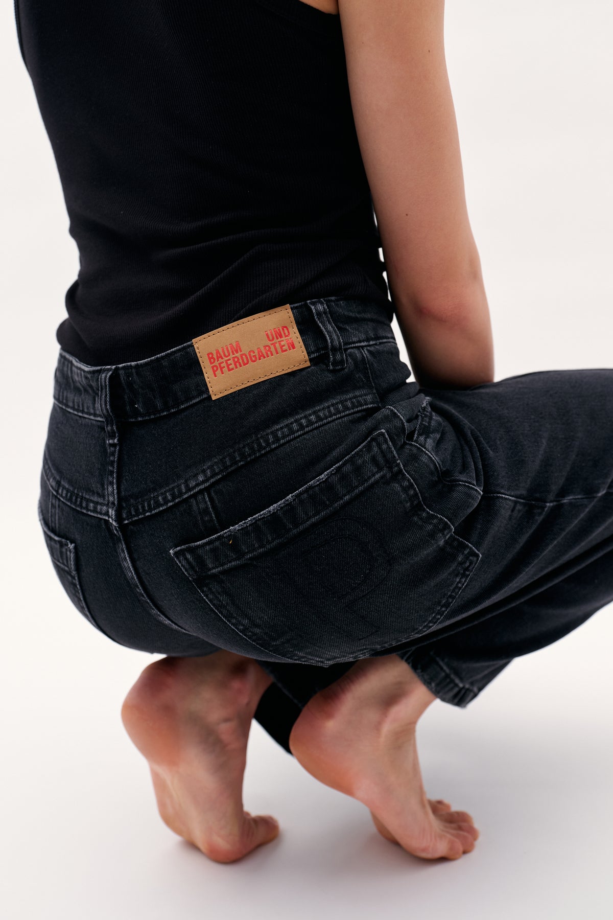 Close-up of charcoal-black jeans’ back pocket and tan Baum und Pferdgarten waistband patch, worn with a black ribbed top.