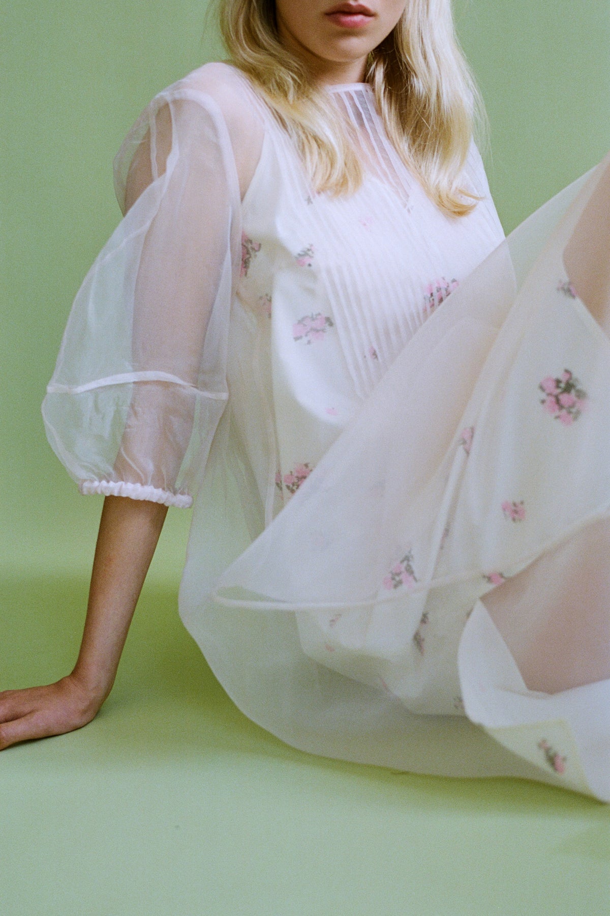 Cropped campaign portrait of a model in a flowing white dress with pink floral details and sheer puff sleeves against a pale green backdrop