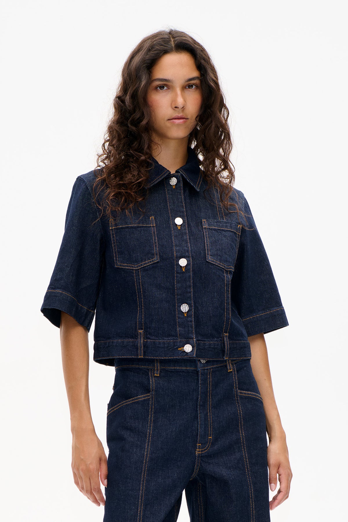 Front view of a dark indigo denim jacket with short sleeves, two chest pockets, silver buttons and contrasting topstitching.