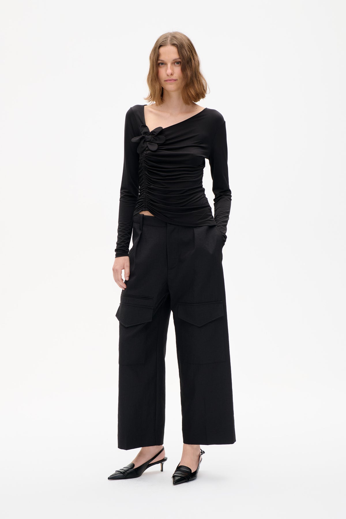Front view of a model wearing a black long-sleeved top with an asymmetric neckline, ruched front and flower detail, paired with wide-leg trousers.