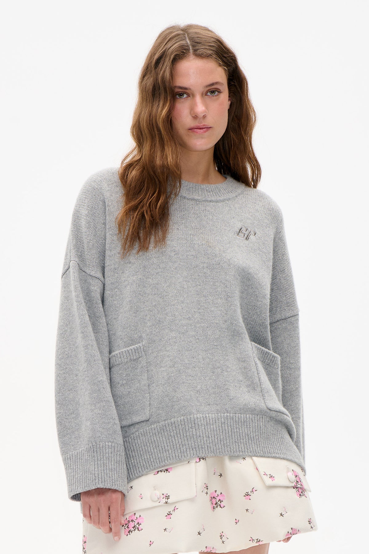 Front view of a light grey knit sweater with ribbed trim and a small chest emblem, worn over a cream skirt with pink floral print.