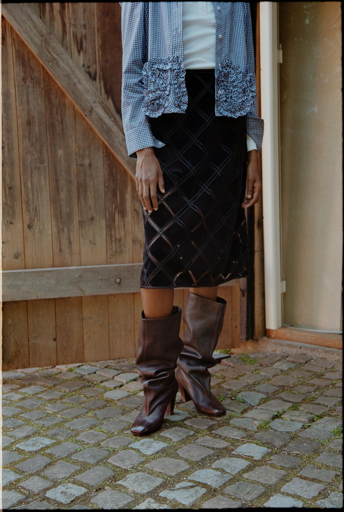 Cropped campaign view of a model wearing an open blue-and-white gingham shirt with ruffled cuffs, a black plaid skirt and tall brown boots.