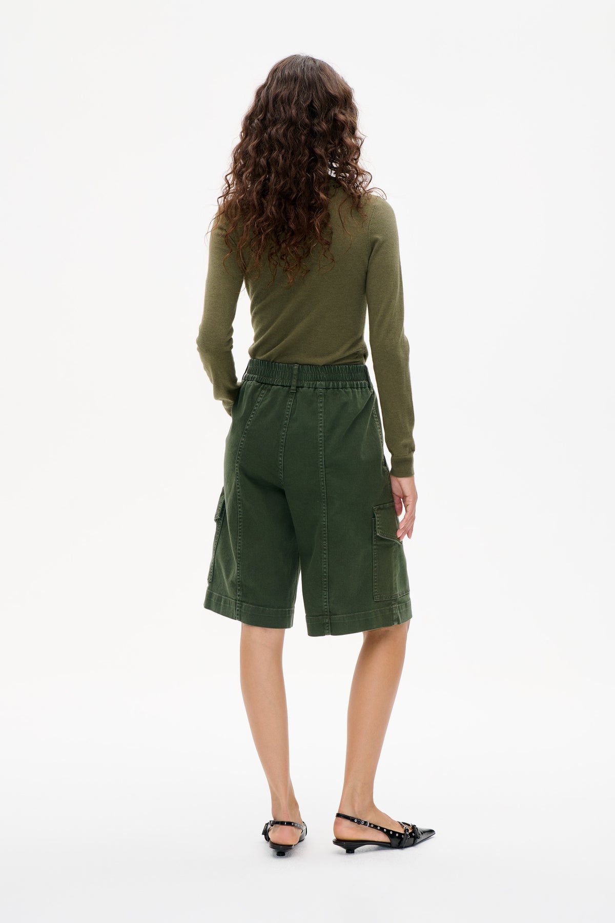 Back view of a model wearing an olive long-sleeved top, dark green cargo shorts with an elastic waistband, and black slingback flats.