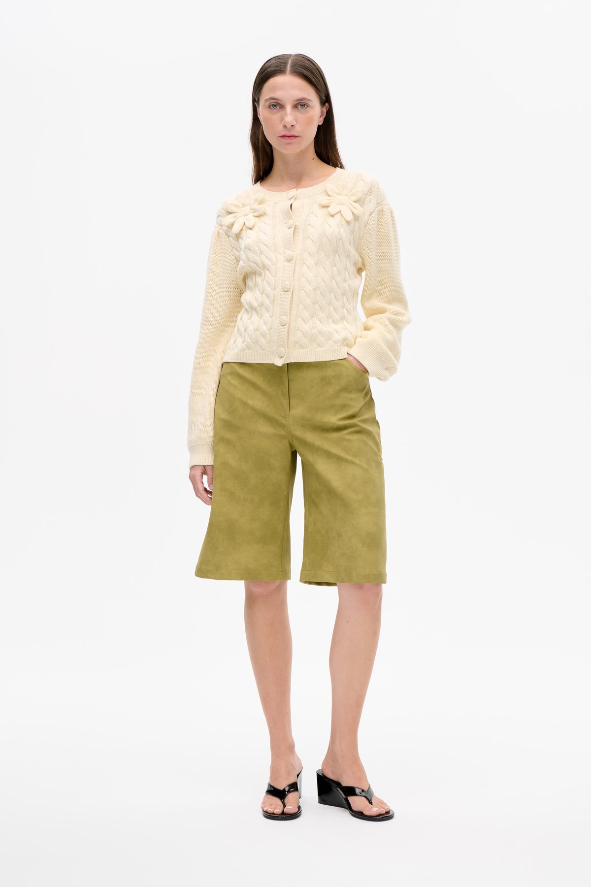 Front view of a model wearing an ivory cable-knit button cardigan with raised flower appliqués and olive-green knee-length shorts.