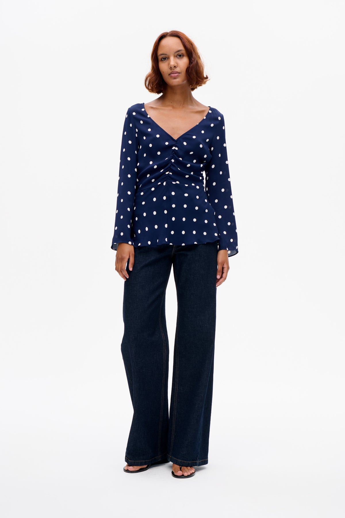 Front view of a navy long-sleeved blouse with white polka dots, a deep V-neck and gathered front, paired with dark wide-leg jeans.