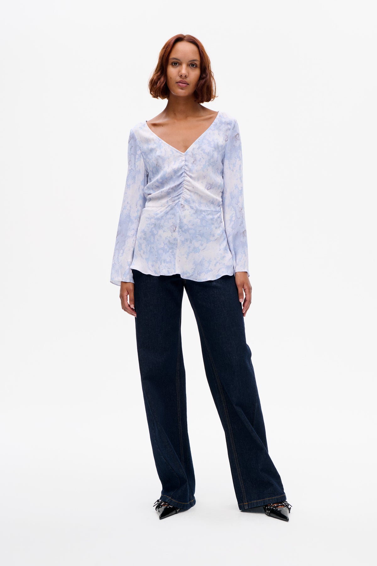 Front view of a model wearing a pale blue abstract floral blouse with a V-neck and gathered front, paired with dark wide-leg jeans.