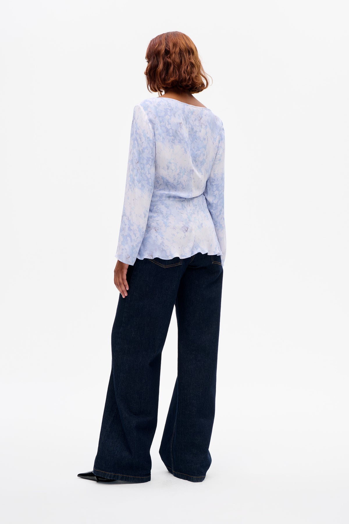 Back view of a model wearing a pale blue floral long-sleeved blouse with a wide neckline and dark wide-leg jeans.