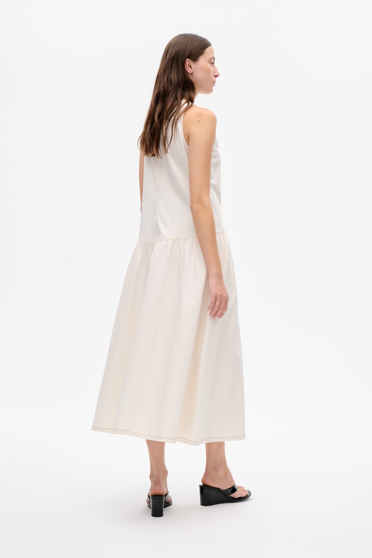 Model in profile wearing an ivory sleeveless midi dress with a gathered skirt and visible contrast stitching.