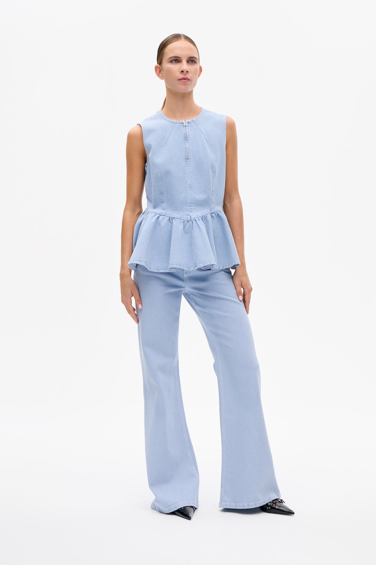 Front view of a model wearing a pale blue sleeveless denim peplum top with a front zip and matching wide-leg jeans.