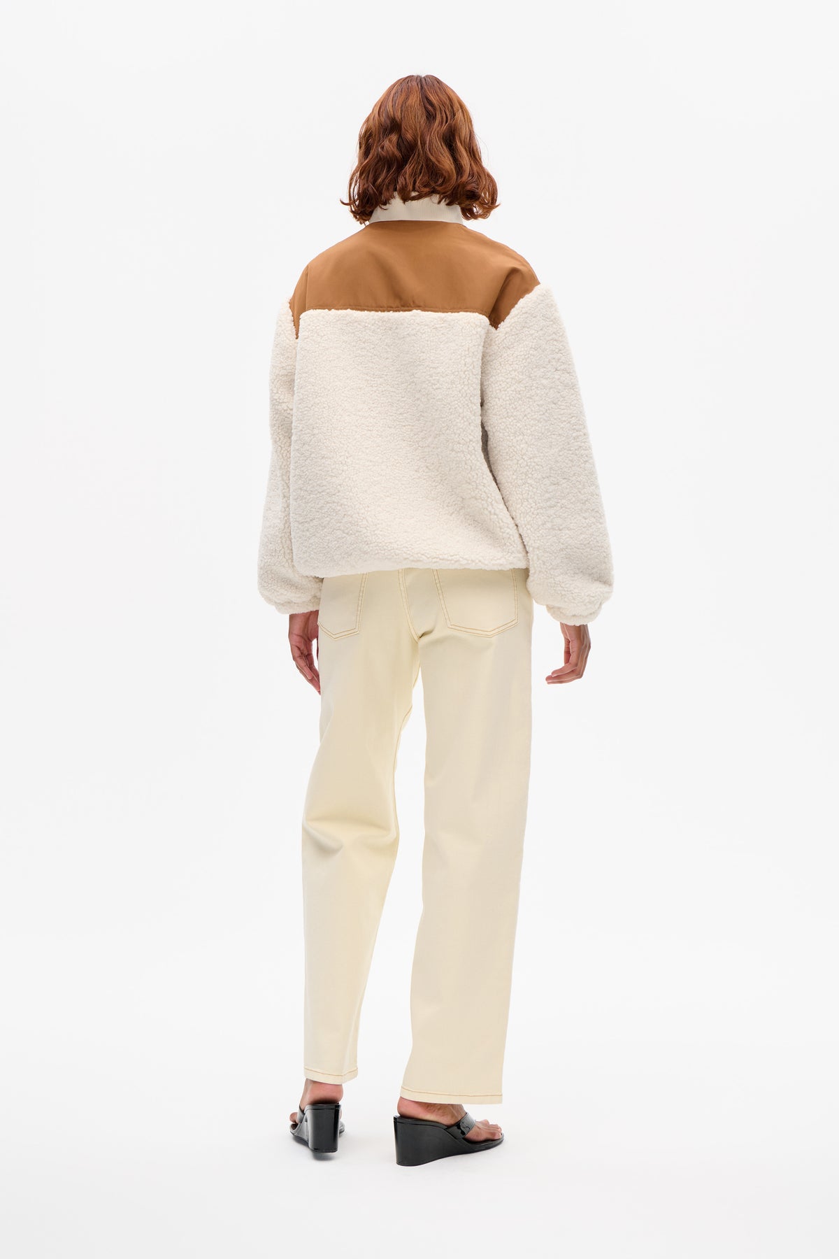 Back view of a cream textured fleece jacket with a camel-brown shoulder panel, styled with pale trousers.