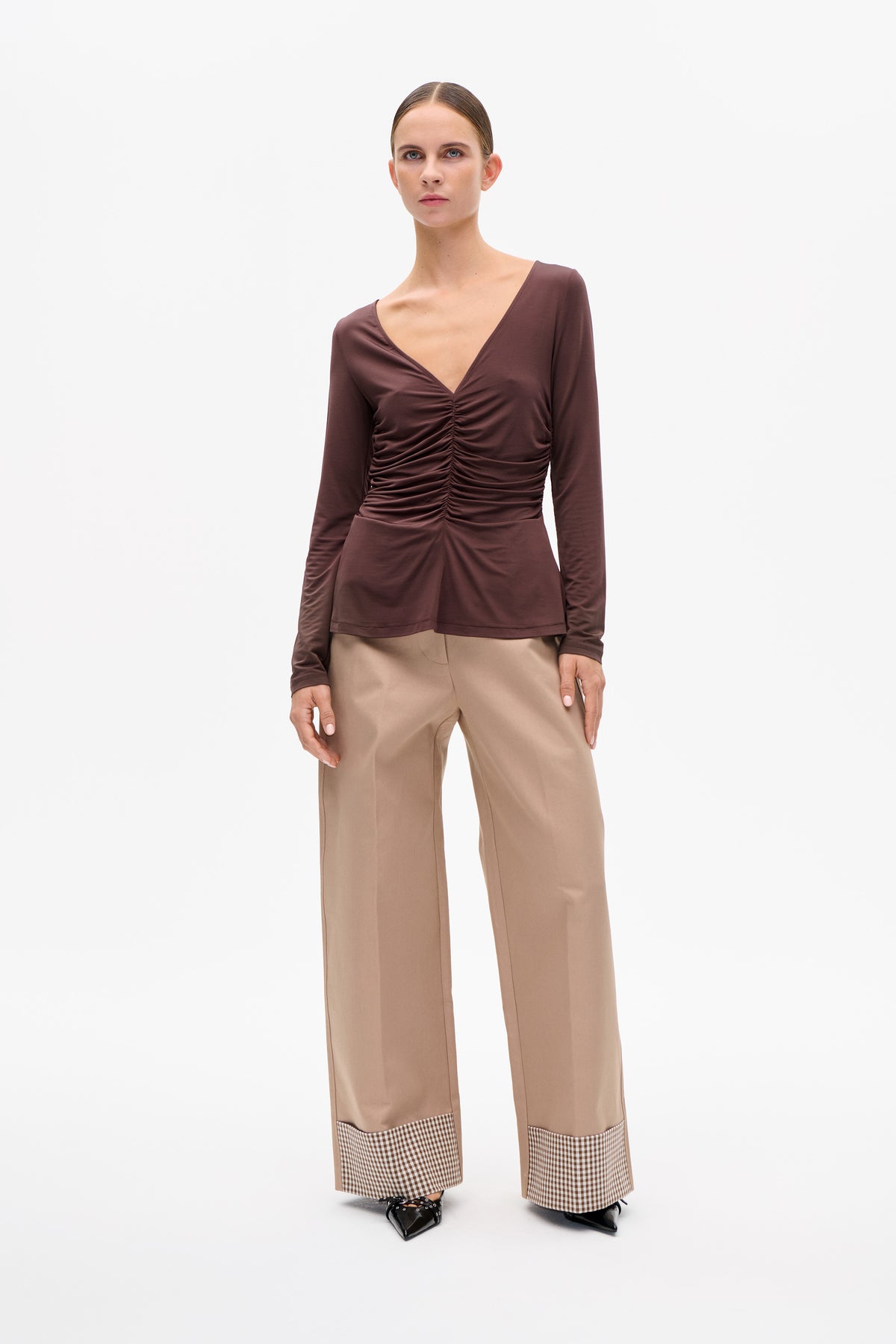 Model wearing a plum long-sleeved V-neck top with a ruched front and beige wide-leg trousers with gingham cuffs.