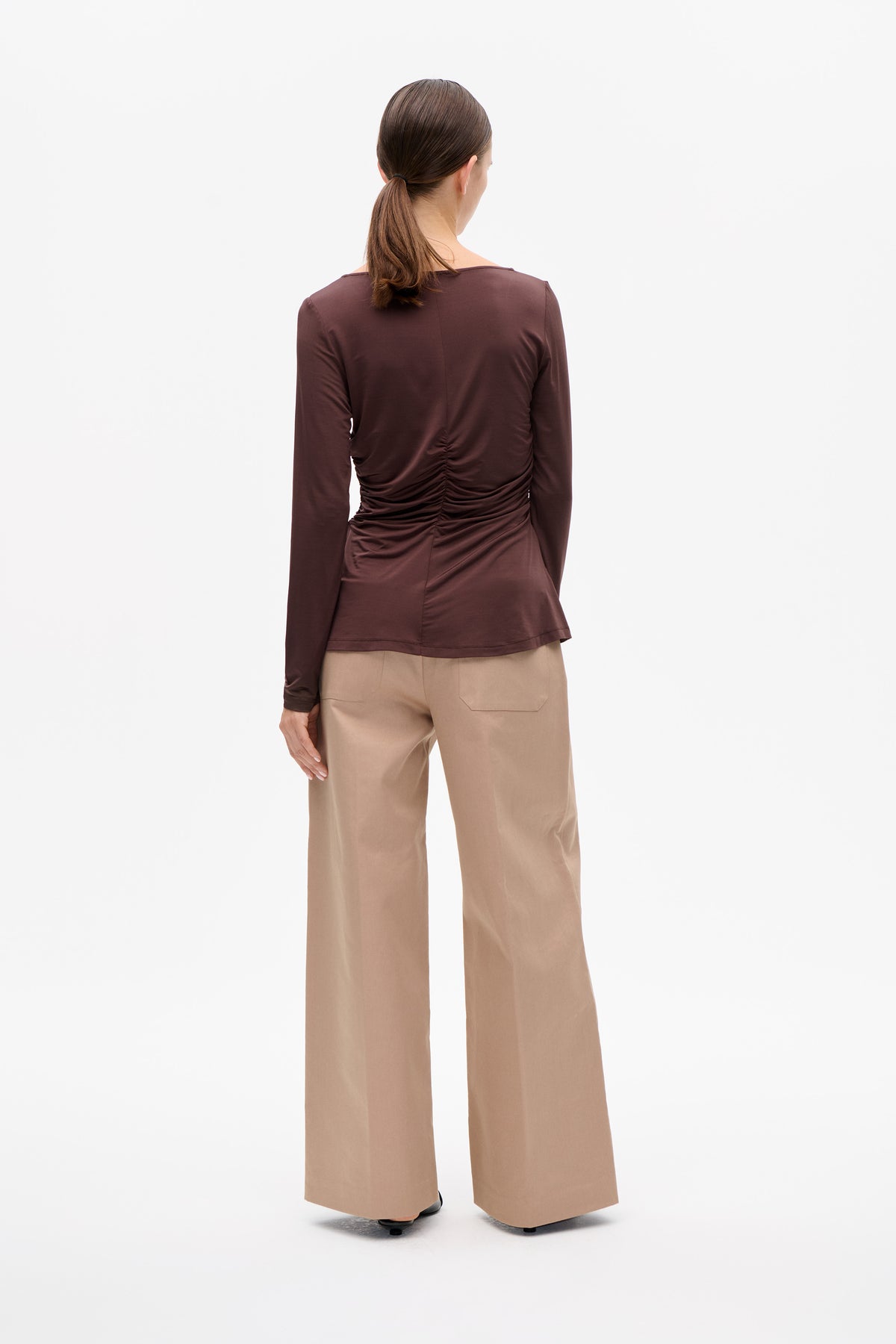 Back view of a model wearing a plum long-sleeved top with gathered ruching down the back and beige wide-leg trousers.