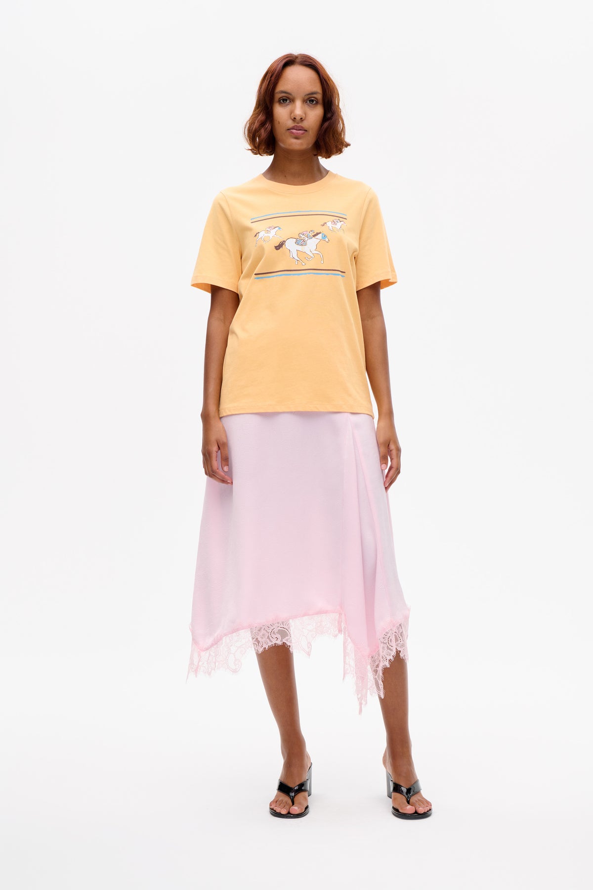 Front view of a model wearing a peach short-sleeve T-shirt with horse graphics and a pale pink skirt with lace trim.