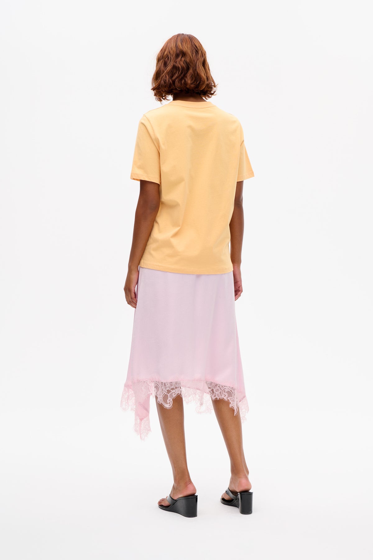 Back view of a model wearing a plain pale yellow short-sleeved T-shirt with a light pink midi skirt featuring a lace-trimmed hem.