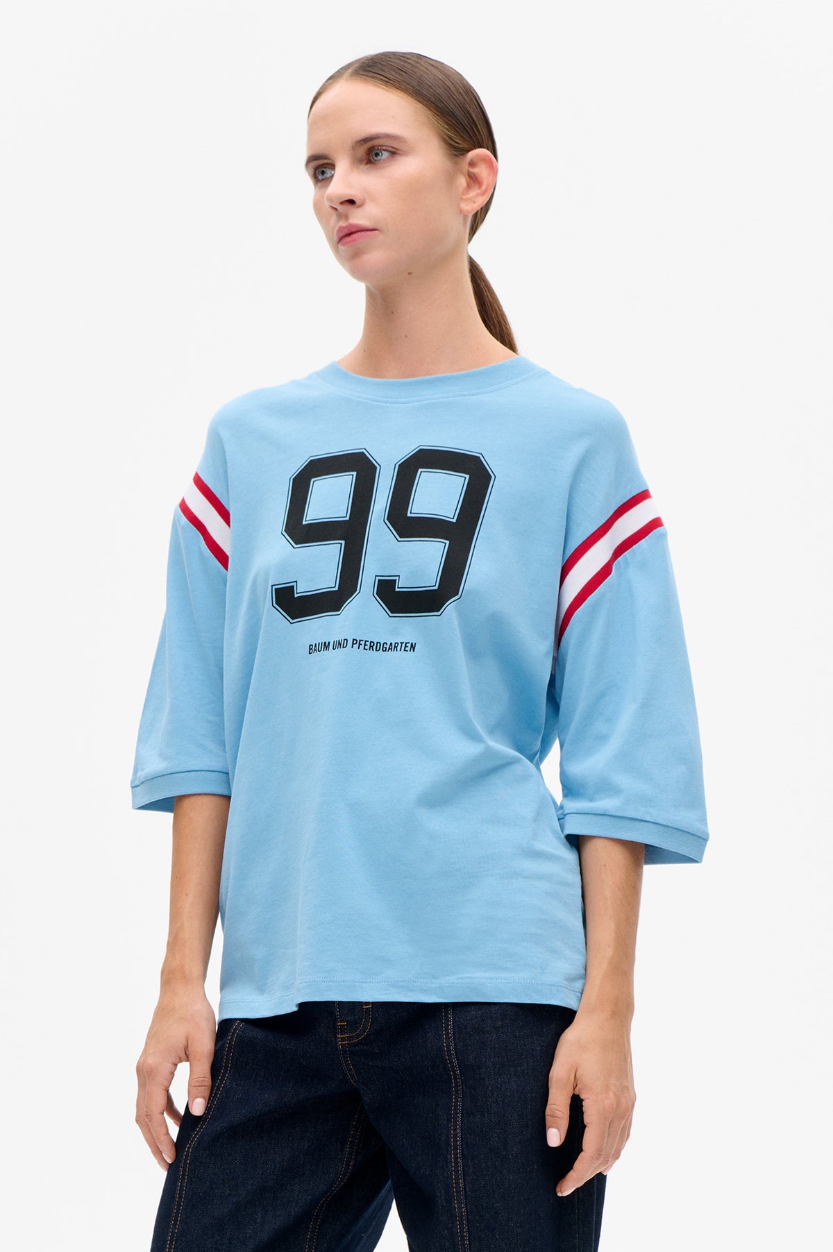 Model wearing a light blue crewneck T-shirt with a large outlined number graphic and red-and-white sleeve stripes, front view.