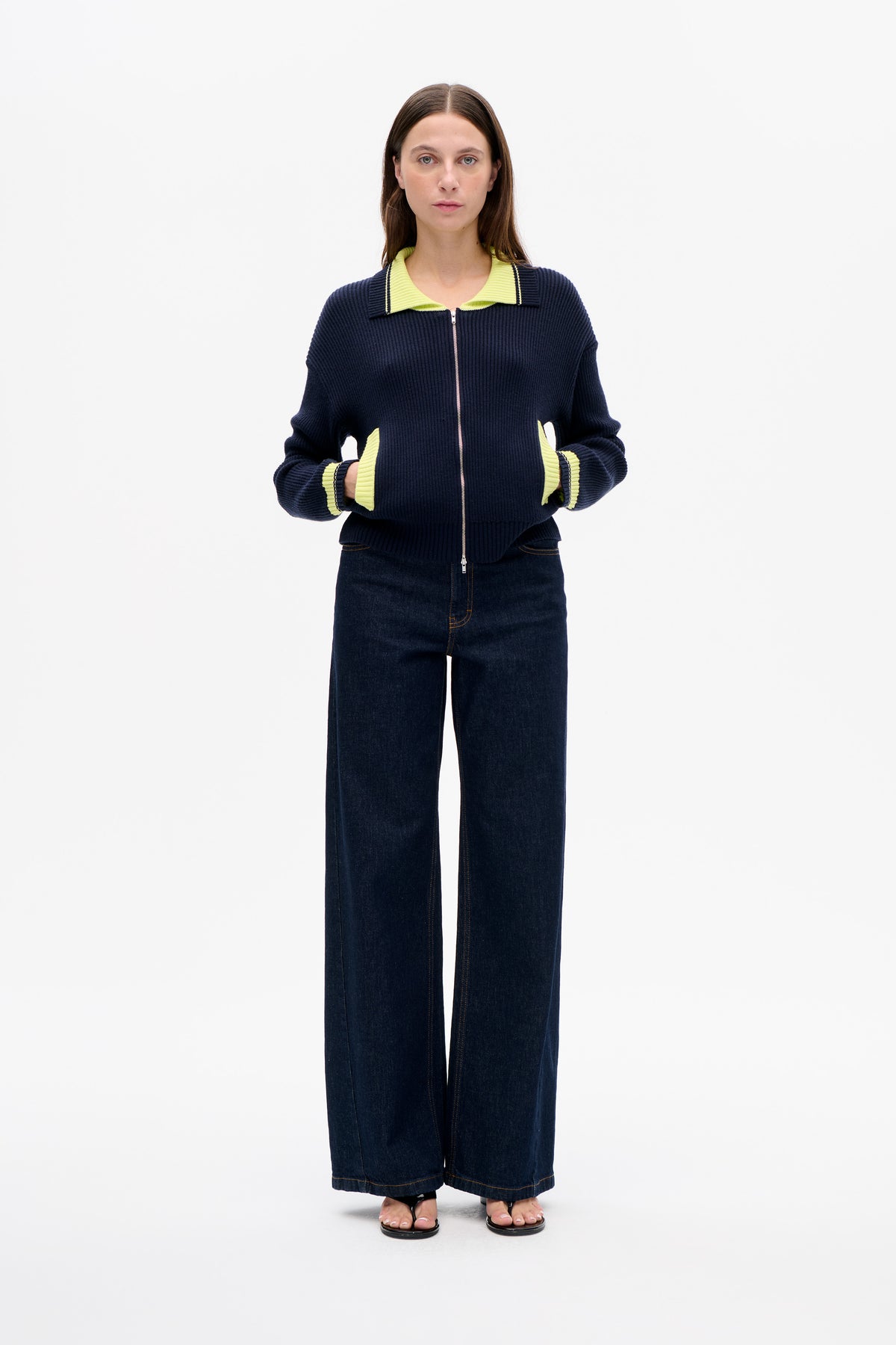 Front view of a model wearing a navy rib-knit zip cardigan with a lime-green collar, pocket trim and cuff stripes, paired with dark wide-leg jeans.
