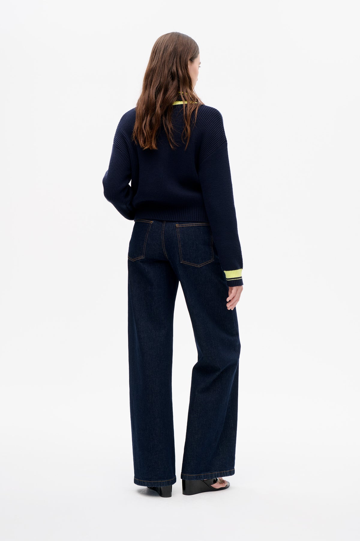 Back view of a model wearing a navy rib-knit sweater with lime accents at the collar and cuffs, paired with dark wide-leg jeans.