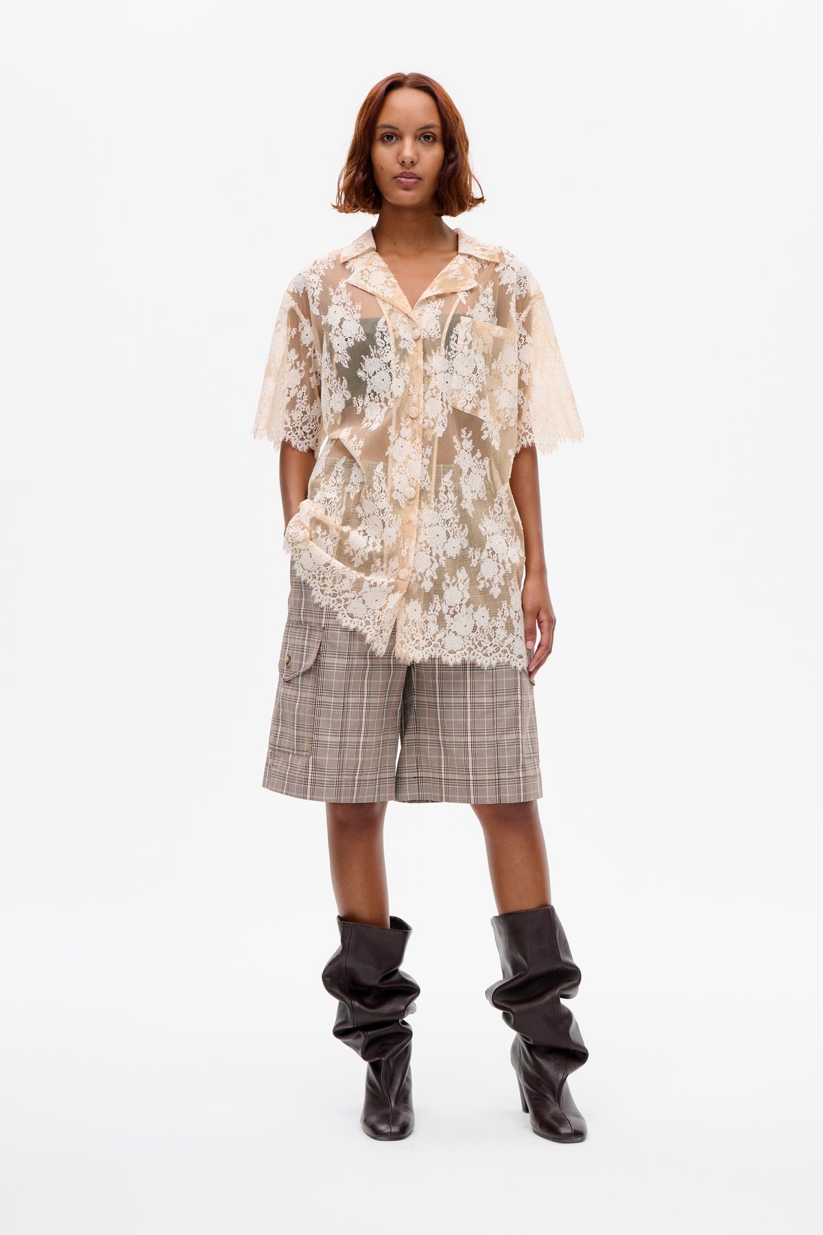Front view of a model in a sheer cream floral lace button-front shirt, beige plaid shorts and dark brown slouch boots.