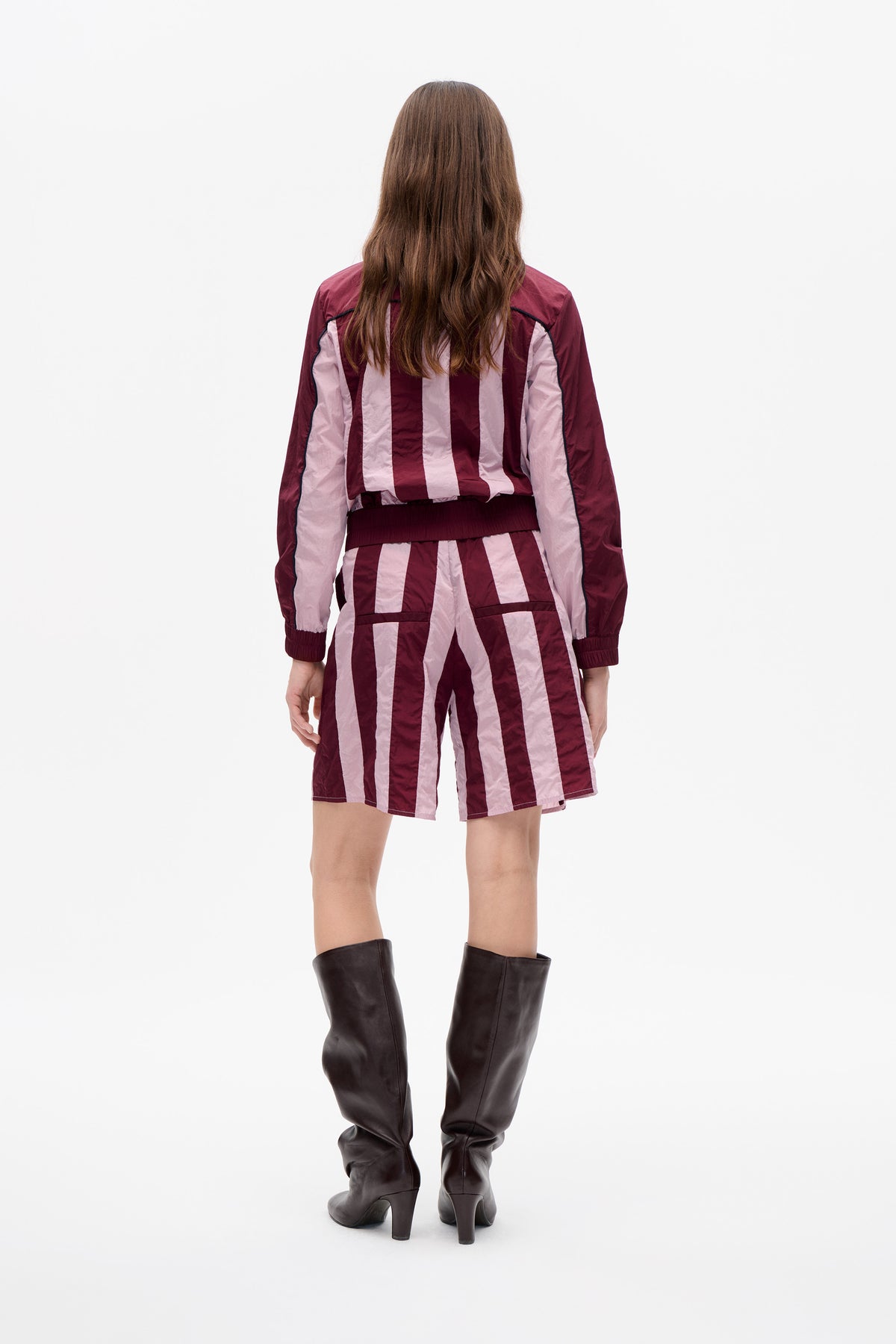 Back view of a model wearing a burgundy and pale pink striped jacket and matching shorts with dark piping and tall dark boots.