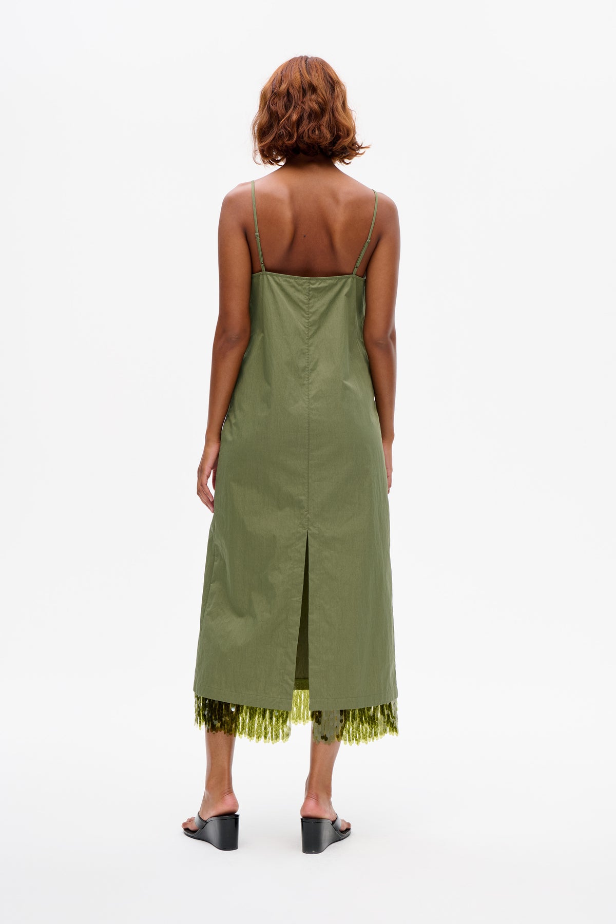 Back view of a model wearing an olive-green strappy midi dress with a center seam and slit, layered over a green sequined skirt.