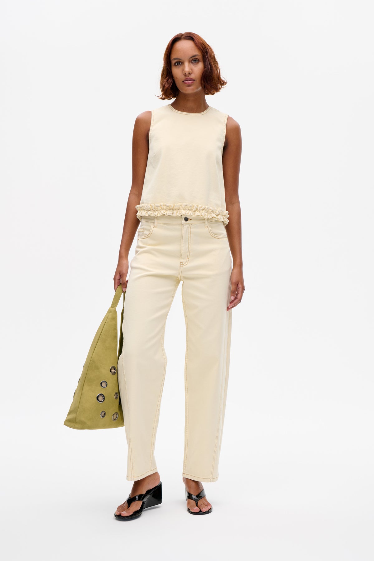 Model wearing a pale cream sleeveless top with contrast stitching and a ruffled hem, paired with matching cropped jeans and an olive tote.