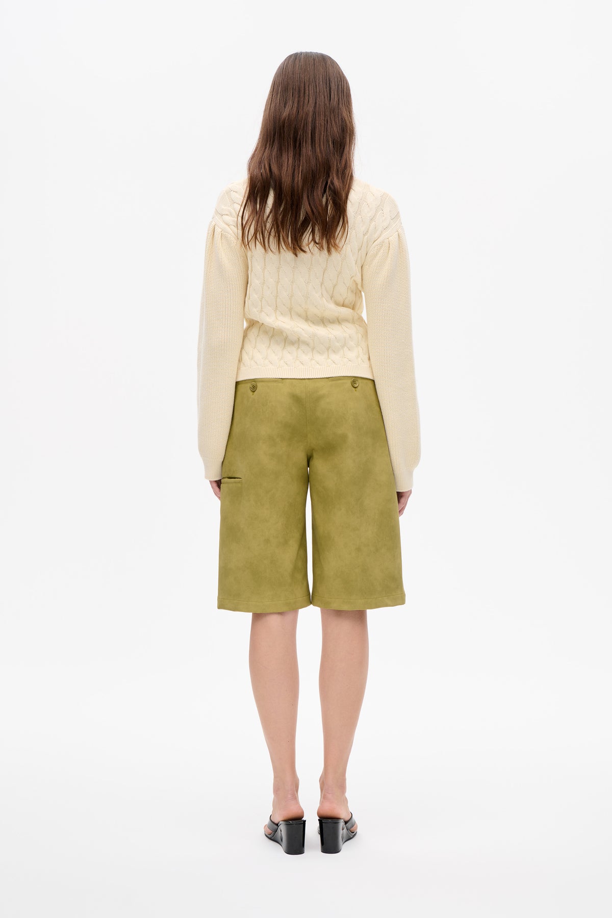 Back view of a model wearing a cream cable-knit sweater with long sleeves and olive-green knee-length shorts.