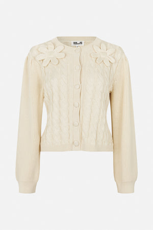 Front view of an ivory cable-knit cardigan with ribbed trim, fabric-covered buttons and large knitted flower appliqués on the shoulders.