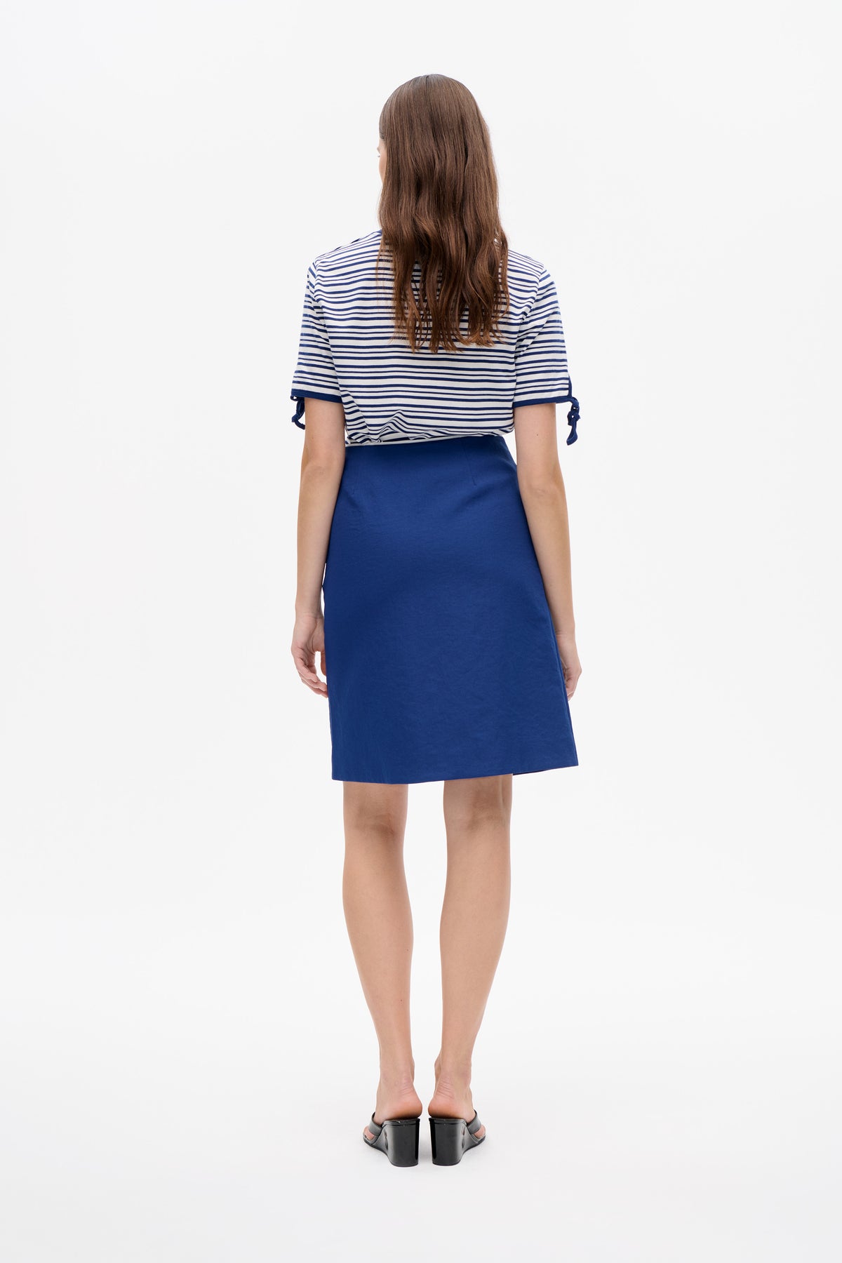 Back view of a model wearing a white T-shirt with navy horizontal stripes and sleeve ties, paired with a solid blue knee-length skirt.