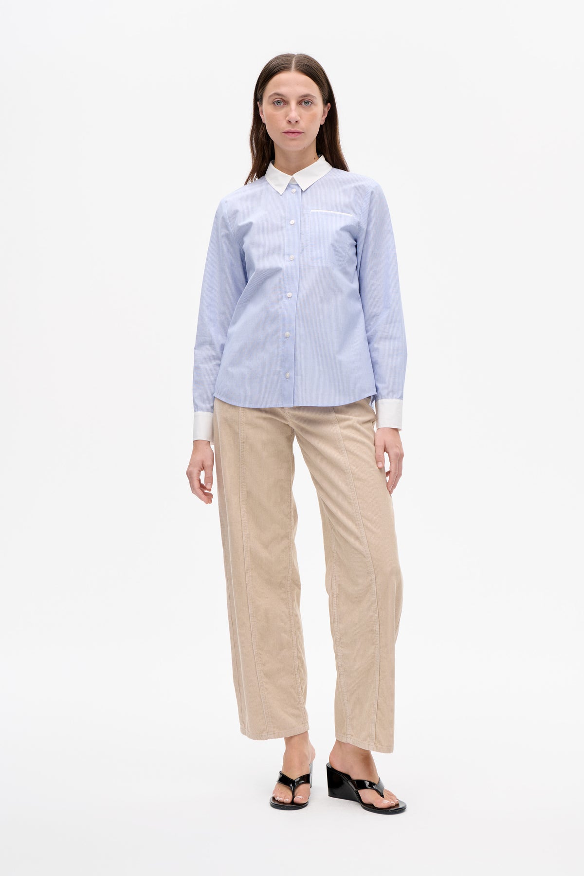 Front view of a model wearing a pale blue fine-striped button-up shirt with a white collar, cuffs and chest pocket, paired with beige cropped trousers.