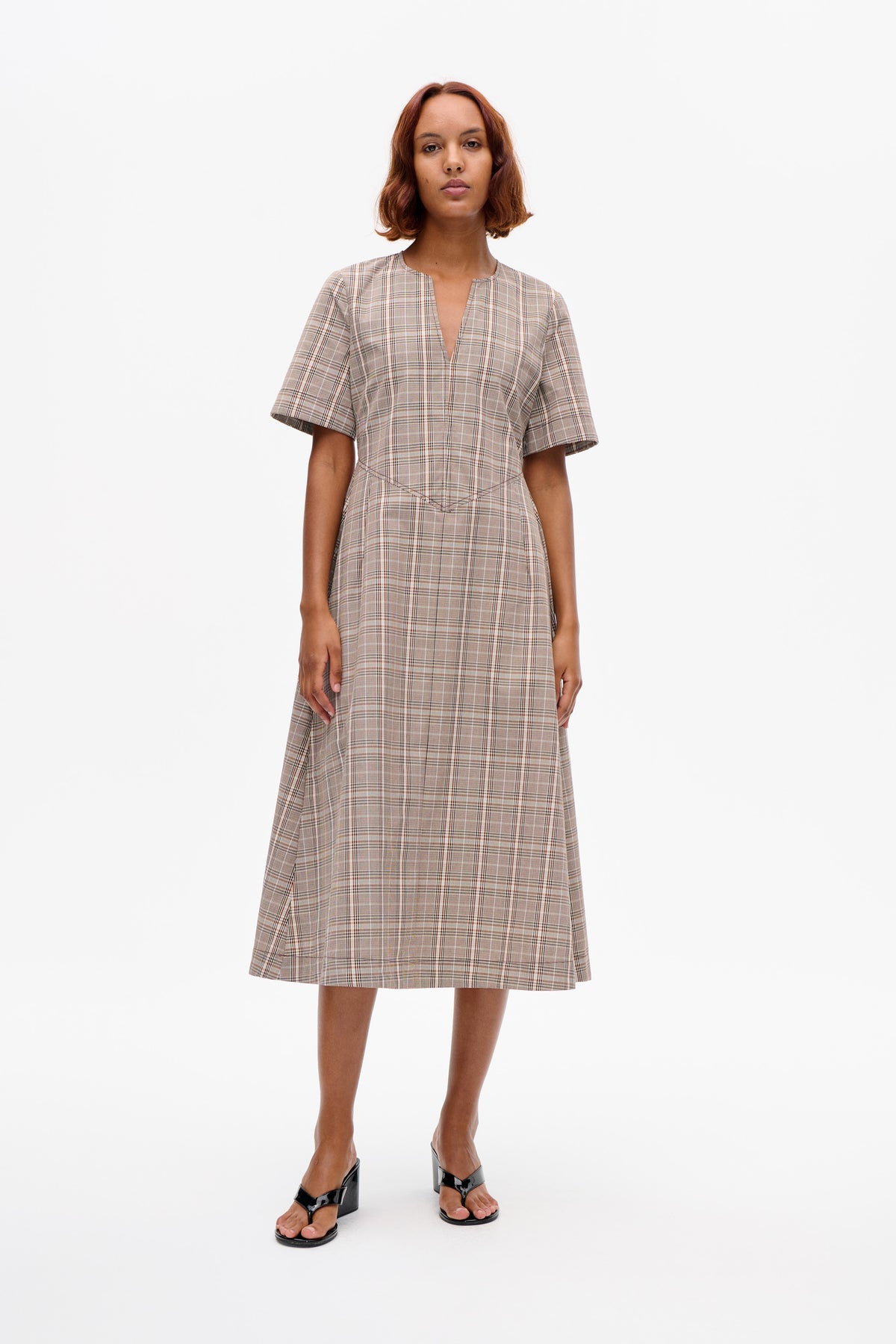 Model wearing a beige and brown plaid short-sleeved dress with a split V neckline and angled waist seams, shown from the front.