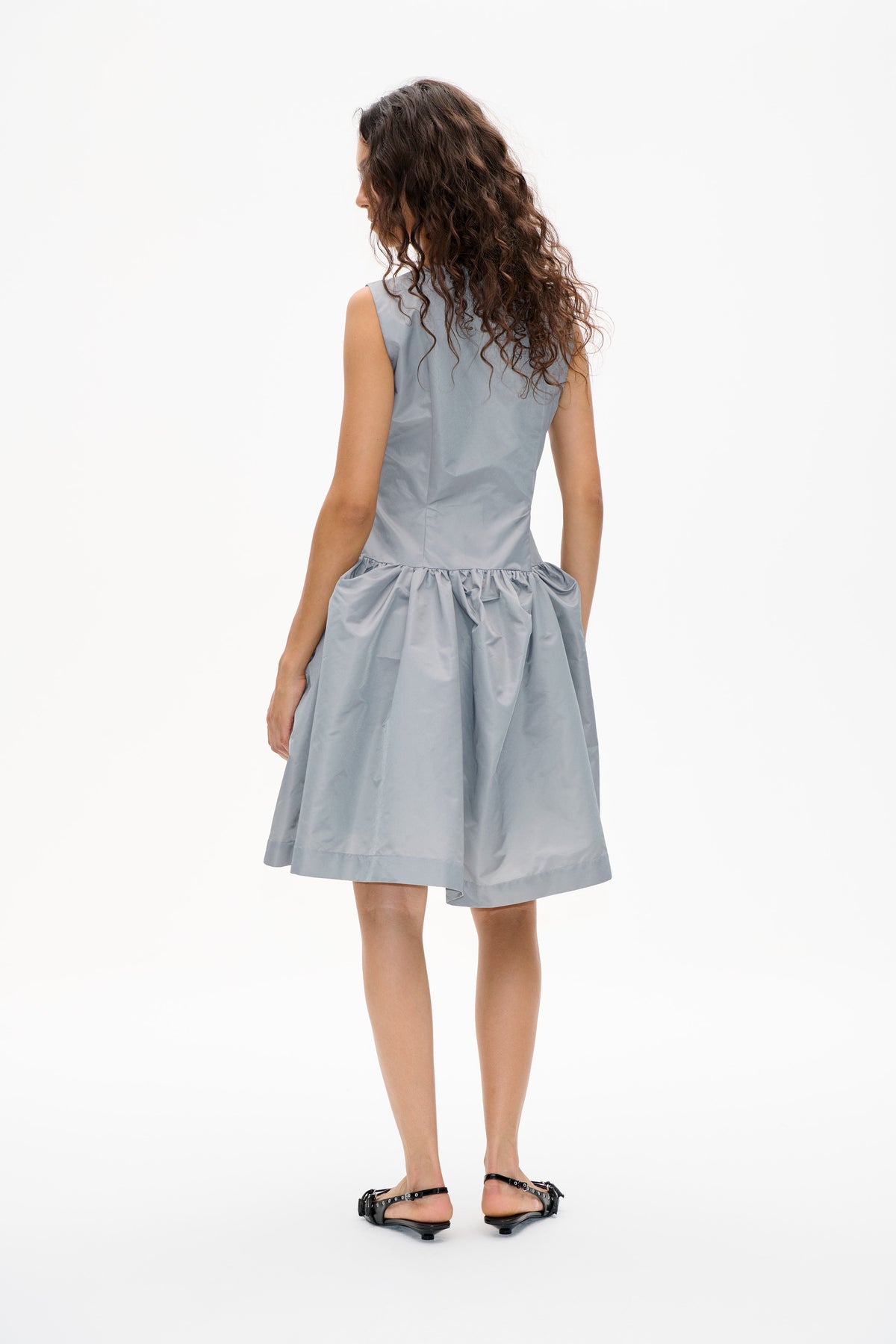 Rückansicht eines Models in einem hellblau-grauen ärmellosen Kleid mit tailliertem Oberteil und gerafftem, voluminösem Rock.