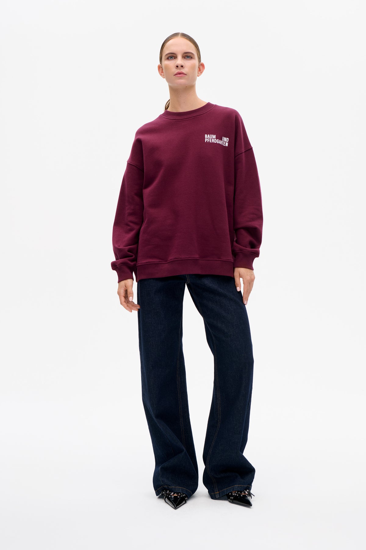 Model wearing a burgundy crew-neck sweatshirt with a small white Baum und Pferdgarten chest logo, dark wide-leg jeans and black pointed shoes.