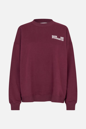 Front view of a burgundy crewneck sweatshirt with ribbed cuffs and hem and white Baum und Pferdgarten lettering on the chest.