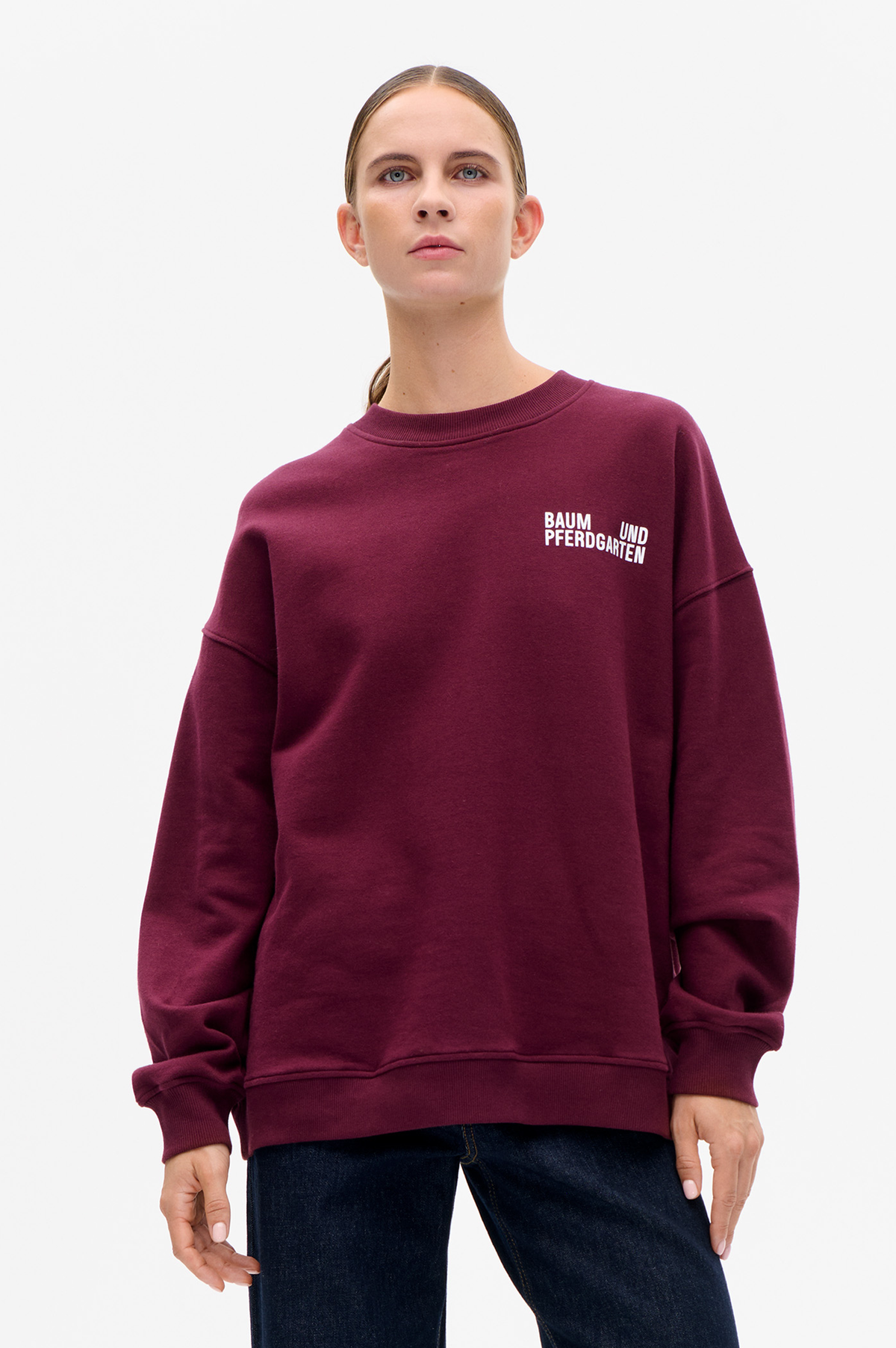 Model wearing a burgundy crew-neck sweatshirt with ribbed trims and white Baum und Pferdgarten chest lettering, shown from the front.