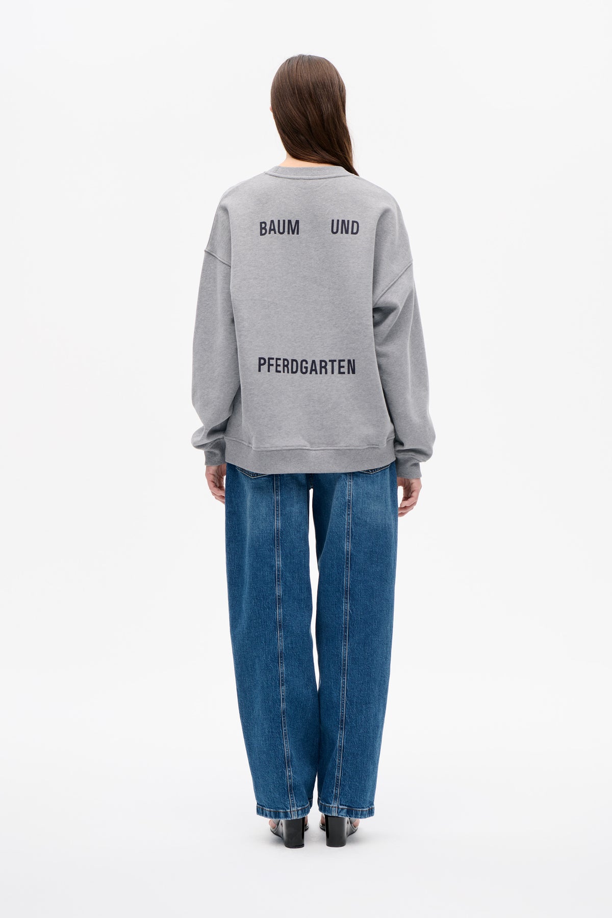 Back view of a model wearing a light grey sweatshirt with dark Baum und Pferdgarten lettering and blue jeans.