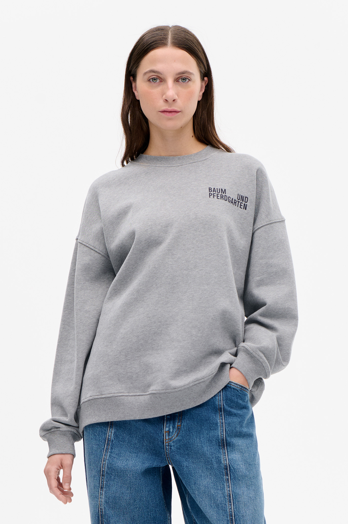 Model wearing a grey crew-neck sweatshirt with black Baum und Pferdgarten chest lettering and blue jeans, shown from the front.