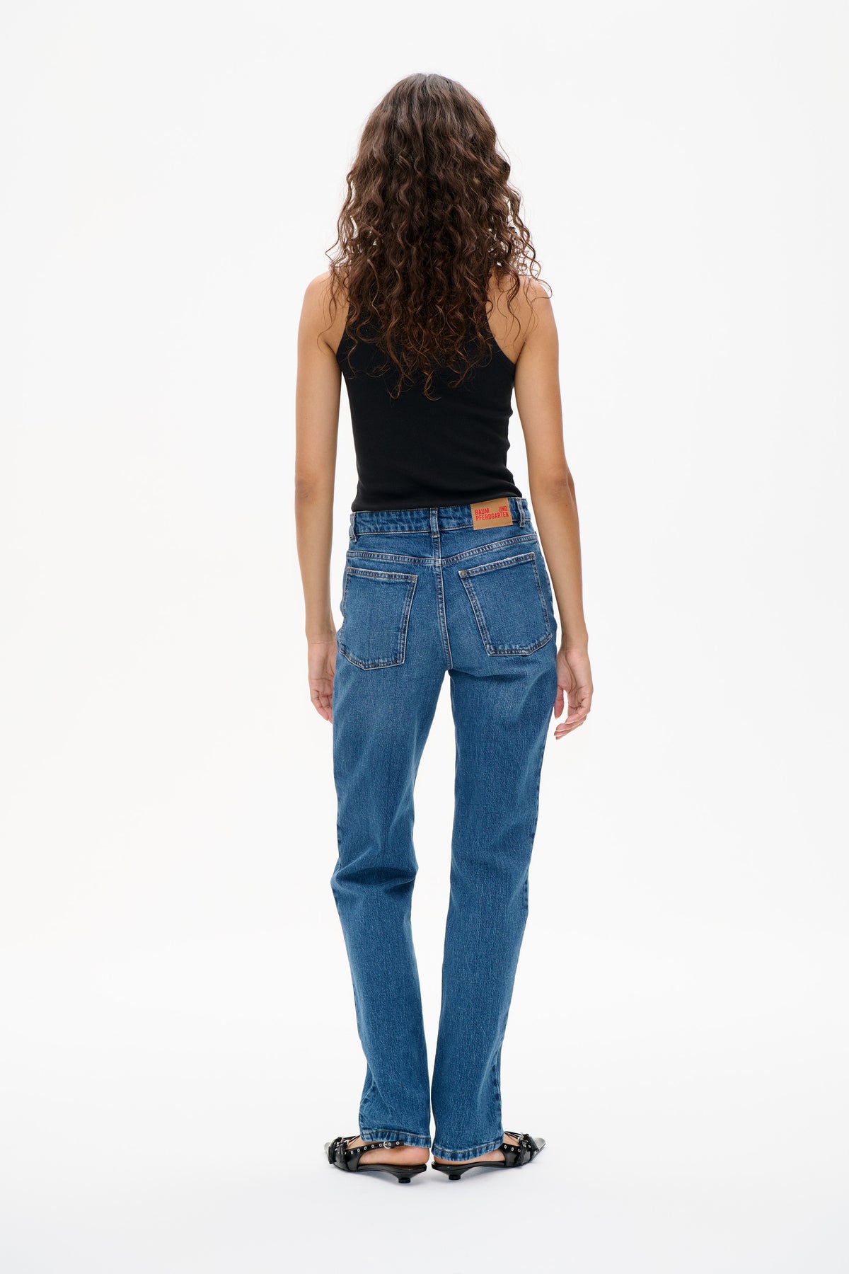 Back view of a model wearing a black sleeveless top, blue straight-leg jeans with rear patch pockets, and black strappy shoes.