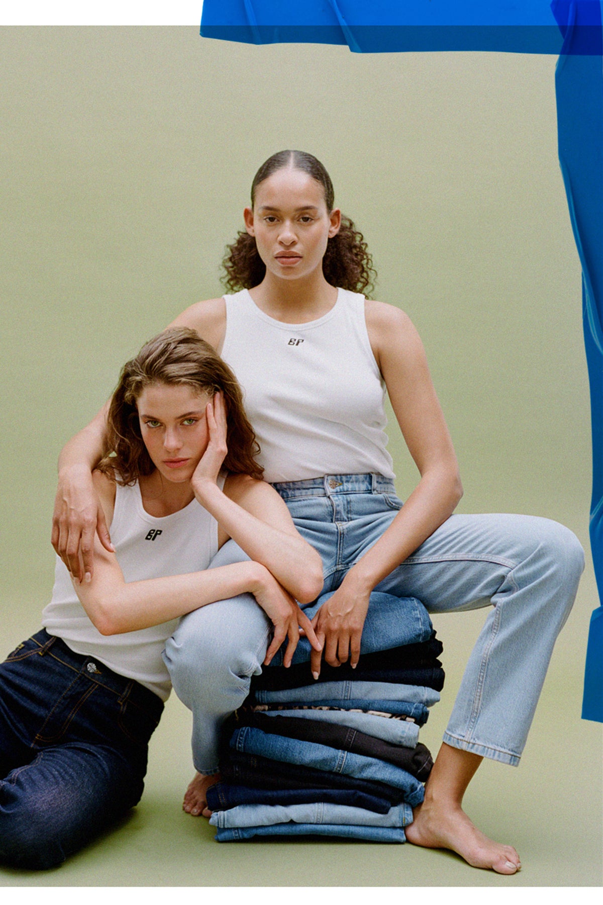 Two models pose in white sleeveless tops and blue jeans beside a stack of folded denim, with a small BP logo on each top.