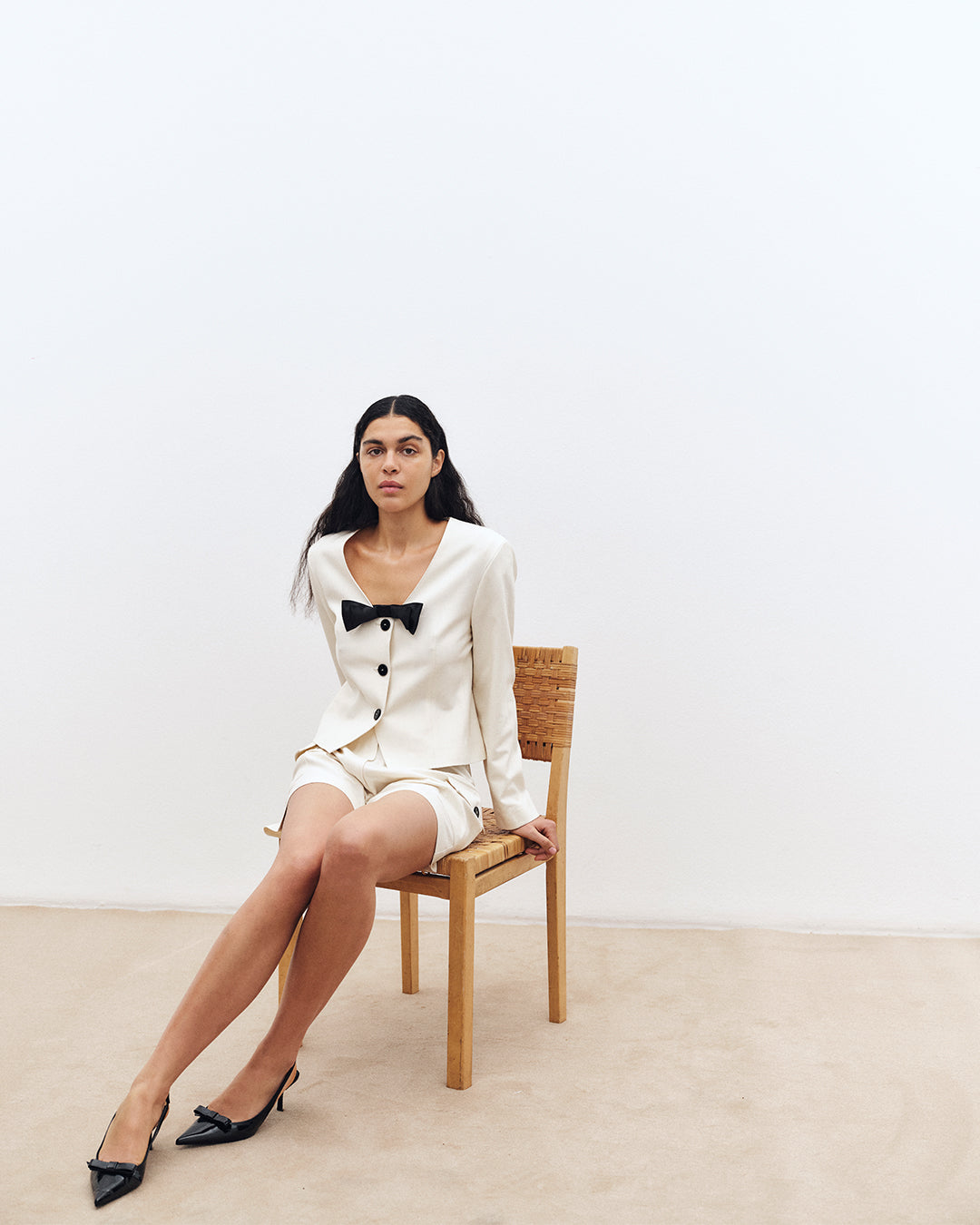 Ein sitzendes Model trägt eine elfenbeinfarbene Jacke mit Knopfleiste und schwarzer Schleife, dazu passende Shorts und schwarze spitze Pumps.